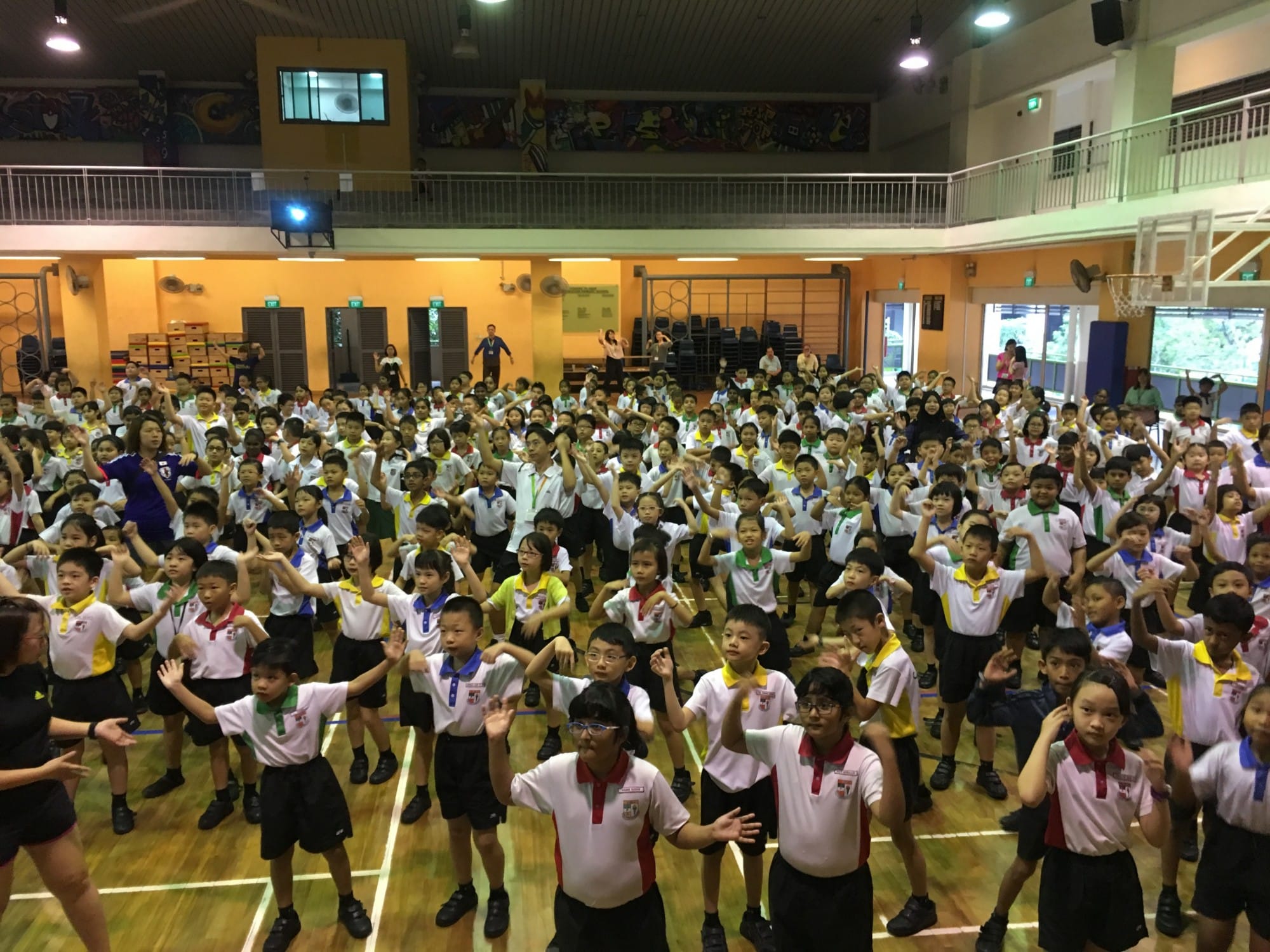 Hundreds of young students in uniforms raise their arms during a group activity inside a school hall.
